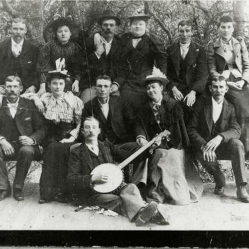 [Portrait of thirteen family members and friends with banjo player at a park]