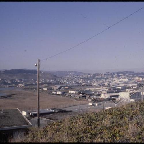 View of fenced off hillside with overgrown vegetation overlooking buildings and warehouses
