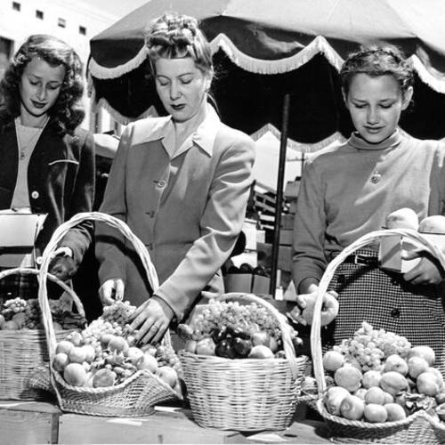 [Three students from the California School of Fine Arts arranging fruit in baskets at the Farmers' Market at Duboce and Market streets]