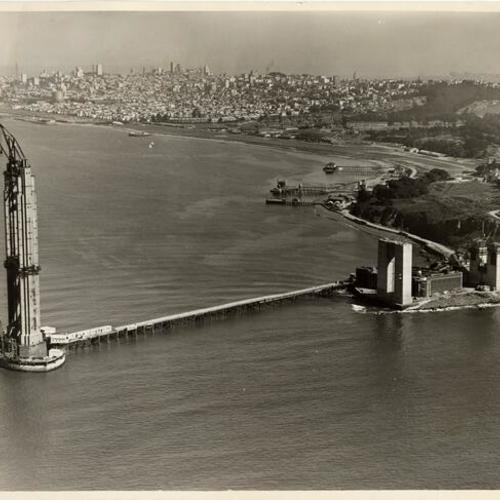 [Aerial view of construction of south tower of Golden Gate Bridge with San Francisco skyline in background]