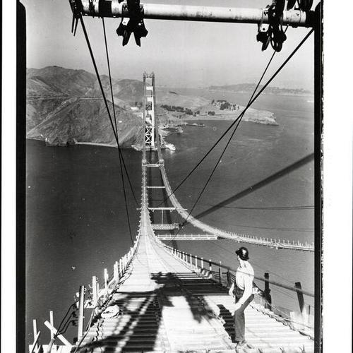 [Workman standing on catwalk construction of Golden Gate Bridge]