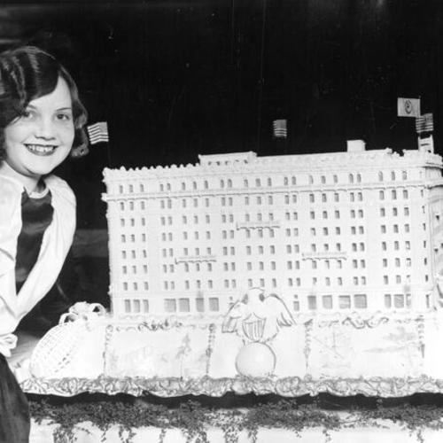 [Young girl standing next to a cake in the shape of the Whitcomb Hotel]