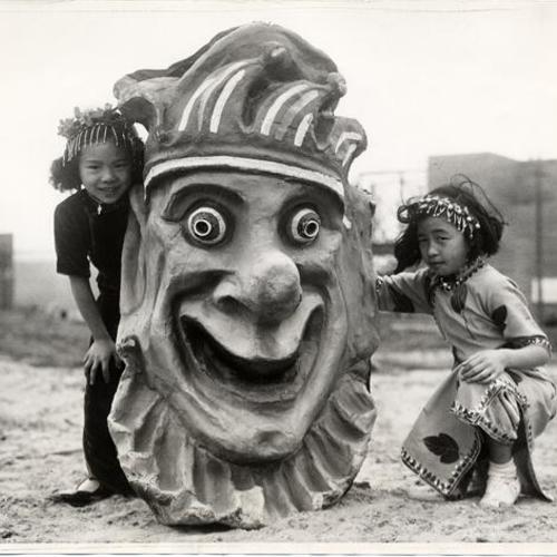 [June and Doris Wong kneeling before a very large mask featured in the 1939 Golden Gate International Exposition]