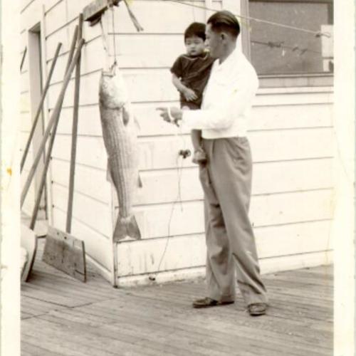 [Unidentified man with a small child and a large fish on a porch in Visitacion Valley]