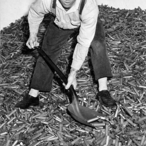[Unidentified worker standing in a room filled with discarded artillery shell casings at the U. S. Mint in San Francisco]