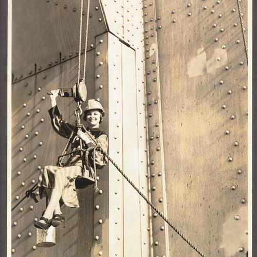 [Woman dangling off the bridge, holding up a paint brush in celebrating opening of the San Francisco-Oakland Bay Bridge]