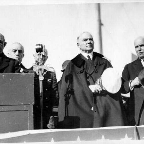 [Officials standing in speakers' stand during groundbreaking ceremony for construction of Golden Gate Bridge while Archbishop Edward J. Hanna delivering invocation]
