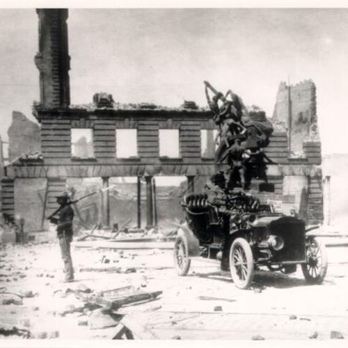 [Soldier standing guard in downtown San Francisco after the 1906 earthquake and fire, with a man sitting in an automobile nearby and the Mechanics Monument in the background]
