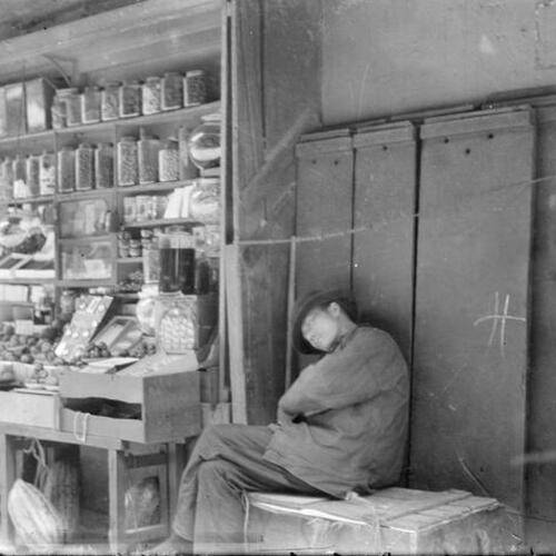 [Clerk sitting, asleep, in open air market. Behind him are fruit and goods (candy?) in jars]