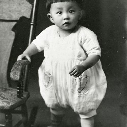[A toddler standing next to wooden rocker in 1925]