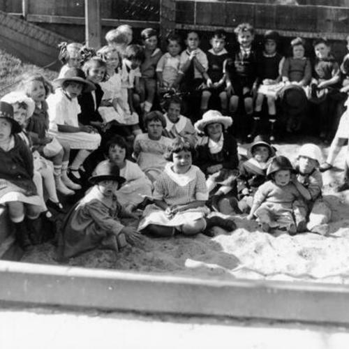 [Unidentified group of children sitting in a sandbox in a playground]