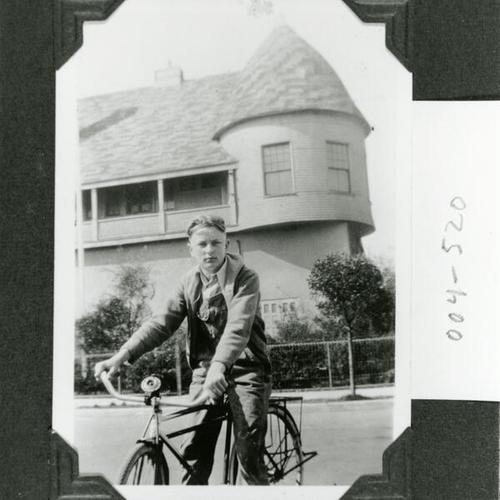 [A young man on a bicycle in front of chalet that was originally located by the beach]