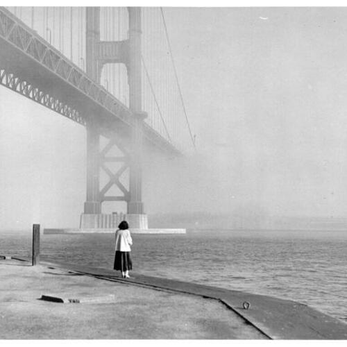 [Unidentified woman standing at Fort Point watching the USS Boxer (obscured by fog) pass underneath the Golden Gate Bridge]
