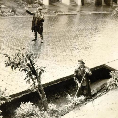 [Man clearing foliage from a trench and another man standing in the middle of the street on Steiner]