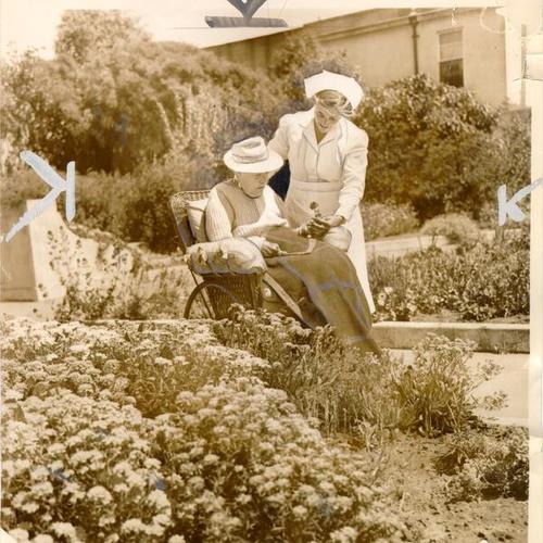 [Nurse Ruth Newbuild wheeling a patient past a garden bed at Garden Nursing Home, 2750 Geary Boulevard]
