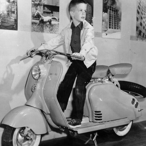 [Tommy Cox standing on a motor scooter during a visit to an "Italy at Work" exhibition at the De Young Museum in Golden Gate Park]