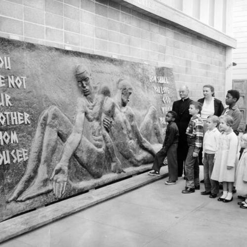 [Father Bruno Drescher, S.V.D. and Mrs. Betty Ford-Aquino standing with a group of children before an 18 foot relief to be placed on the side of the St. Benedict Youth Center]