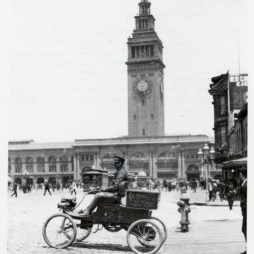 [Lester L. Whitman sitting at the tiller of a Curved Dash Olds Light Runabout in front of the Ferry Building]